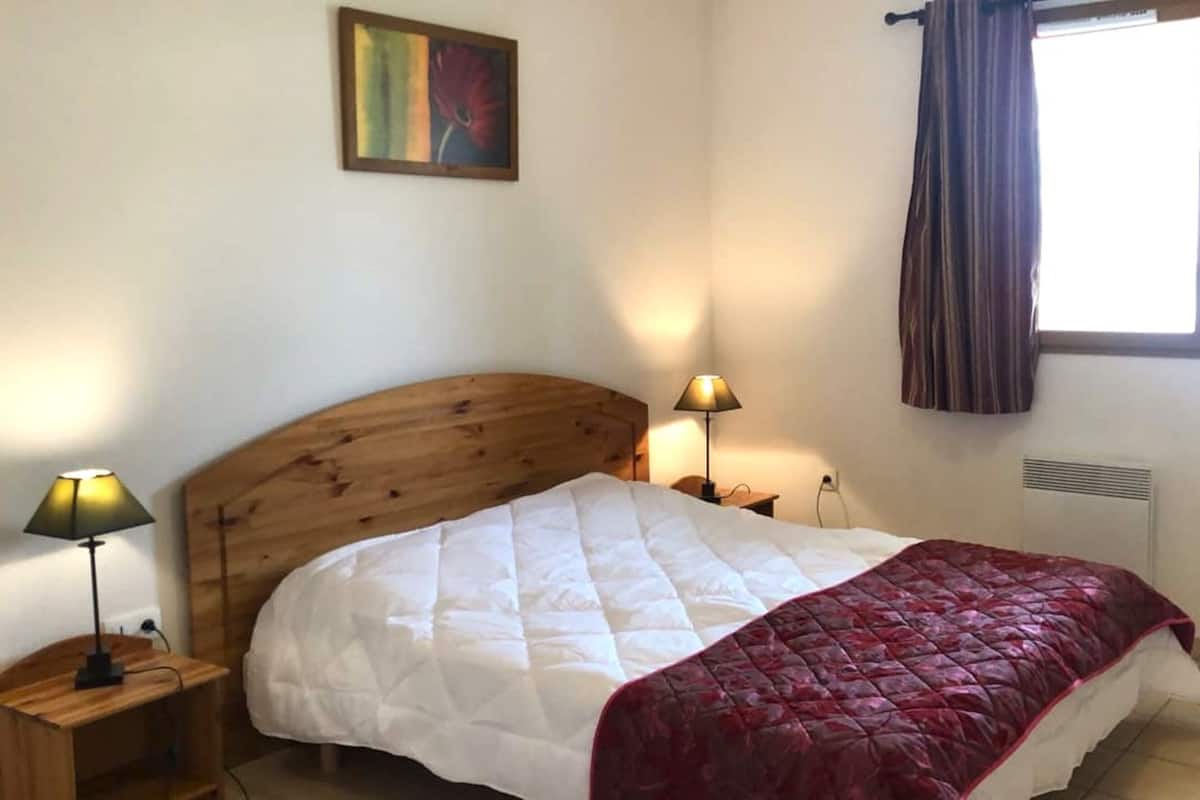 Un dormitorio con cabecera de madera, colcha blanca acolchada, manta roja, mesita de noche con lámpara y ventana con cortinas.