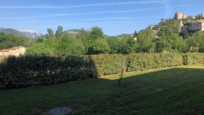 Ein grüner Bereich mit einer Hecke, einem kleinen Baum und einem Gebäude mit einem Türmchen auf einer Anhöhe.