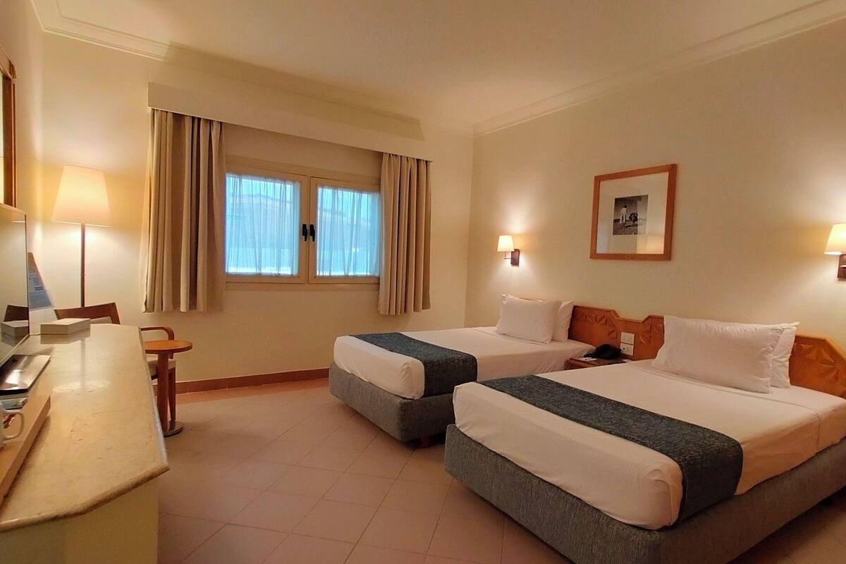 Ein Hotelzimmer mit zwei Betten, einem Schreibtisch mit Stuhl, einem Fernseher und einem Fenster mit Vorhängen.