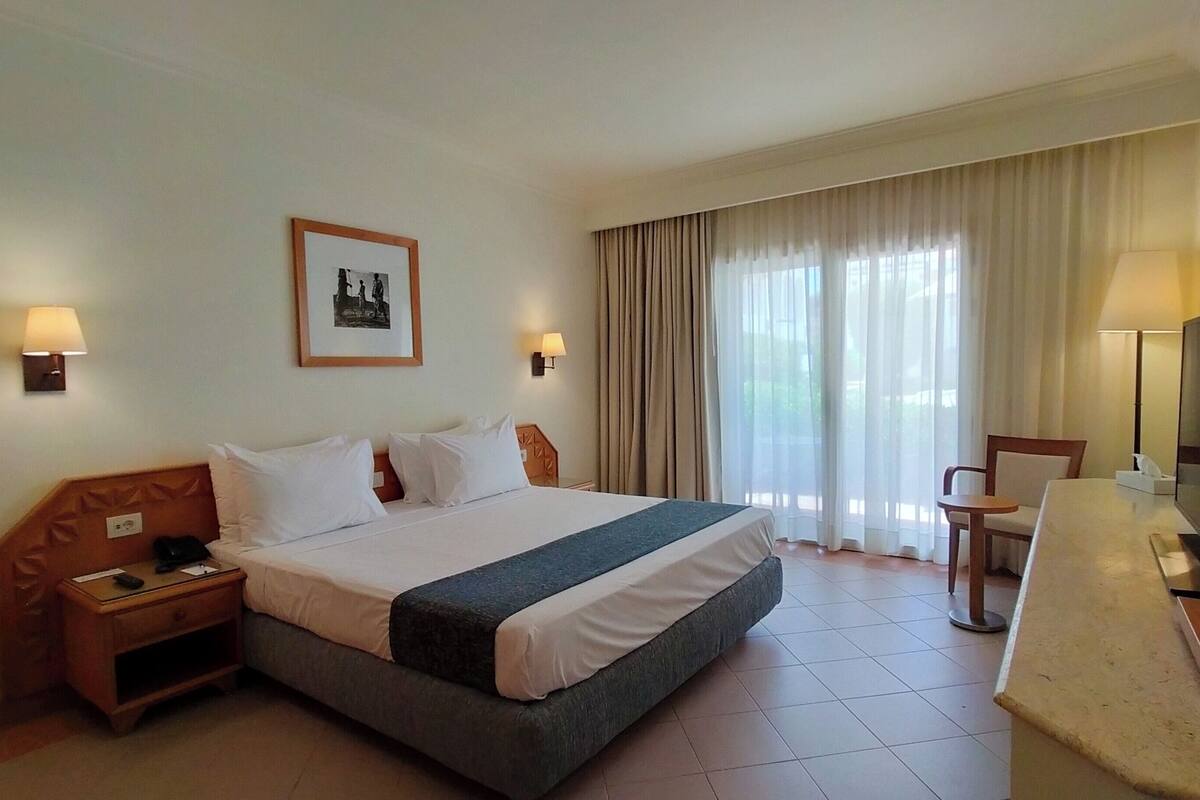 Ein Hotelzimmer mit Bett, Nachttischen, einem Schreibtisch mit Stuhl, einem Fernseher und einem Fenster mit Vorhängen.
