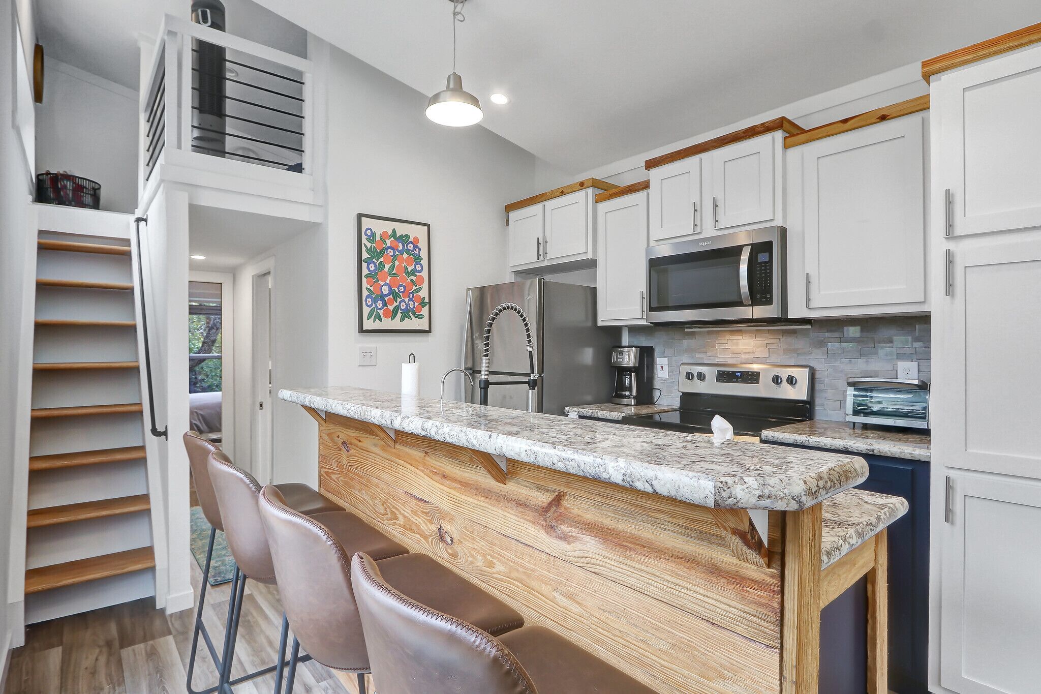 A modern kitchen with white cabinets, a stainless steel refrigerator, a microwave, and a coffee maker. There is a wooden bar counter with bar stools.
