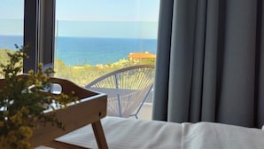 Een slaapkamer met een groot raam met uitzicht op een strand, een houten stoel en een plant op het bed.