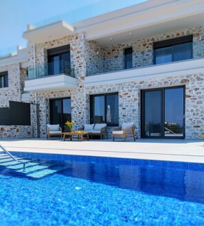 Un edificio moderno con fachada de piedra y ventanas grandes, un área de piscina con sillones de descanso y una piscina azul cristalina.