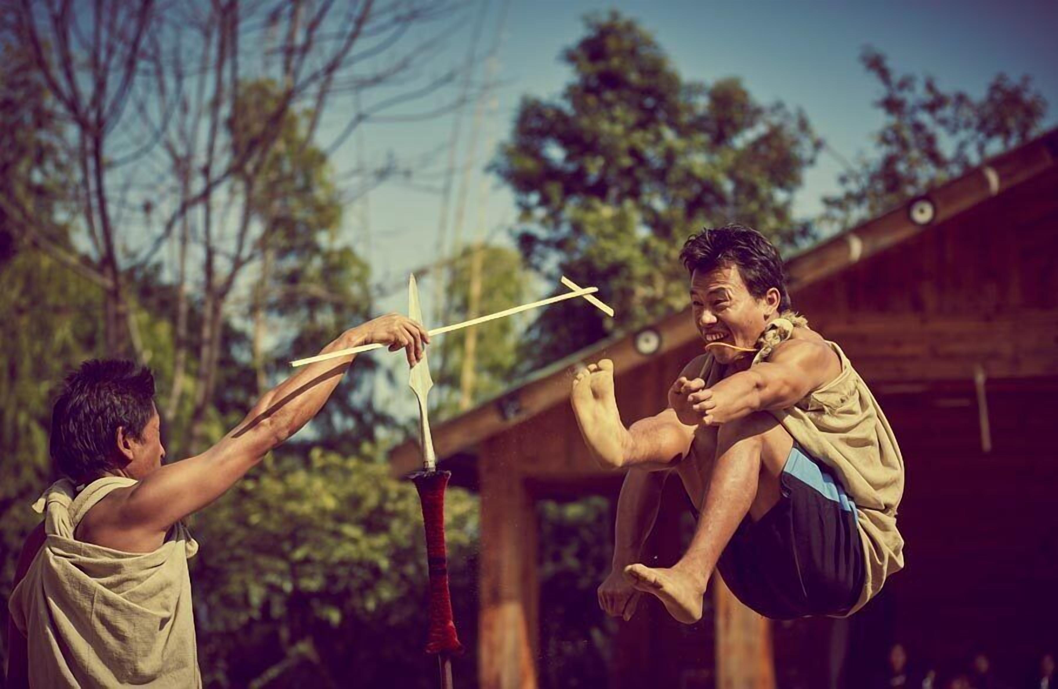 A person performing a martial arts move outdoors with a wooden structure in the background.