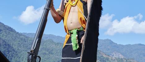 Una persona de pie en una azotea con un rifle, vestida con atuendo tradicional, y un paisaje montañoso al fondo.