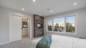 A bedroom with a stone fireplace, a large window with a view, and a neatly made bed. - Luxury Home in Quiet Neighborhood near Downtown with Fast Wi-F & 3 Workspaces (Calgary)