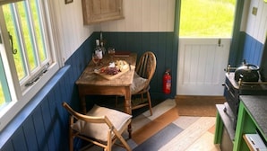Eine gemütliche Küche mit Holztisch für zwei Personen, einem Fenster mit Jalousien, einem Holzschrank und Blick auf eine grüne Rasenfläche.