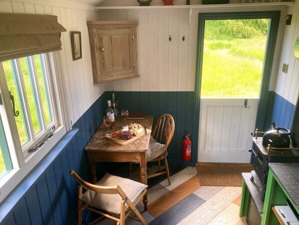 A cozy kitchen with a wooden table set for two, a window with blinds, a wooden cabinet, and a view of a green lawn. - Stompy's hut - Cosy hut in very peaceful location (Laxfield)