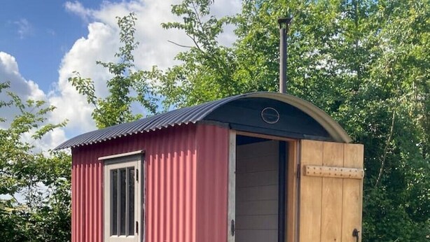 Eine kleine, rote Holzhütte mit einem Fenster und einer Tür, umgeben von viel Grün und Blumen.