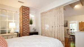 Ein Schlafzimmer mit einem großen Bett, einer Ziegelwand, einem Fenster mit Vorhängen und einer kleinen Küchenzeile.