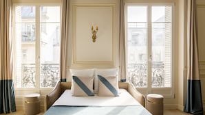 A hotel room with a large bed, two bedside tables, a decorative wall piece, and a view of the city through large windows. - Merveil - Signature Suite - Louvre - Opera I (Paris)