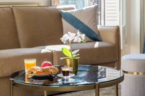 A breakfast tray with pastries, juice, and coffee on a glass table, a potted orchid, and a sofa in the background. - Merveil - Signature Suite - Louvre - Opera I (Paris)