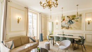 A spacious living room with a beige sofa, a round coffee table, and a dining area with a glass table and chairs. - Merveil - Signature Suite - Louvre - Opera I (Paris)