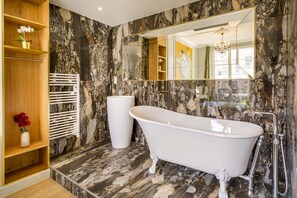 Ein Badezimmer mit freistehender Badewanne, Marmorwänden, einem großen Spiegel und einem Holzregal mit Blumen.