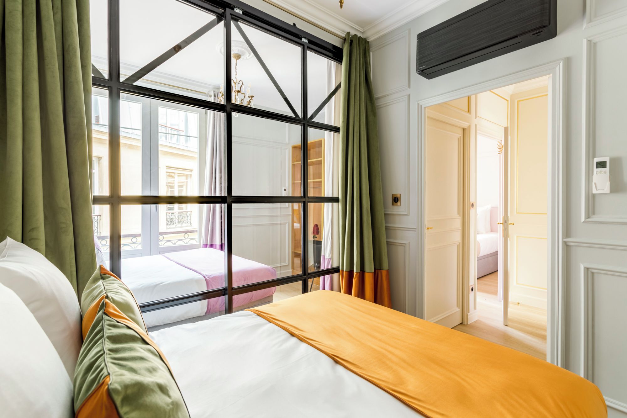 A hotel room with a large window, a bed with a white and orange bedspread, and green curtains.