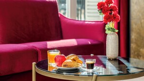 A breakfast tray with croissants, an apple, orange juice, and coffee on a glass table in a room with a red sofa and a vase of red flowers.