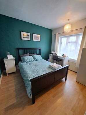 A bedroom with a bed, a nightstand, a radiator, and a window with curtains. - GREEN SWAN (Greenford)