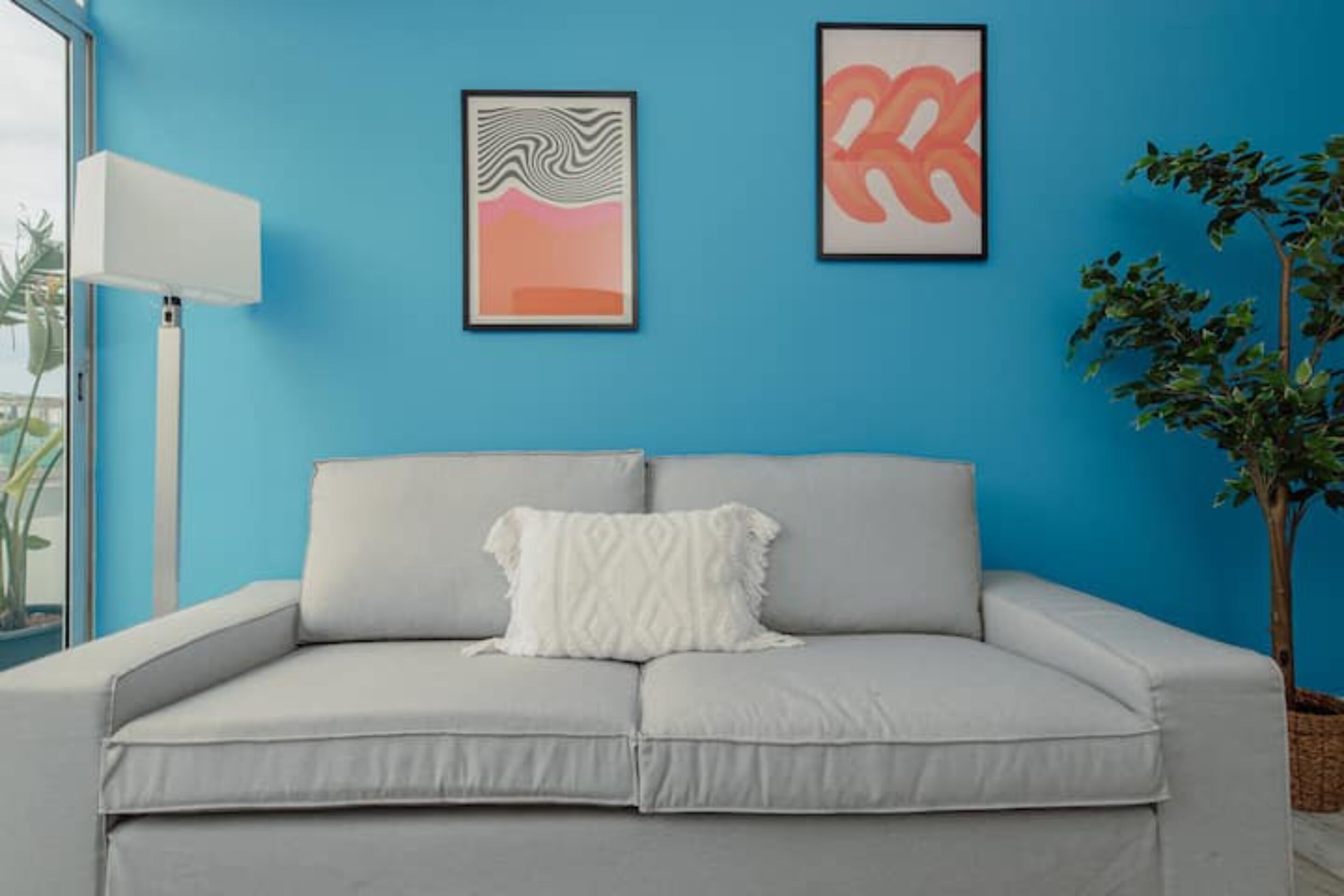 Un canapé gris clair avec un coussin blanc, placé devant un mur bleu et encadré de deux œuvres d’art abstraites.