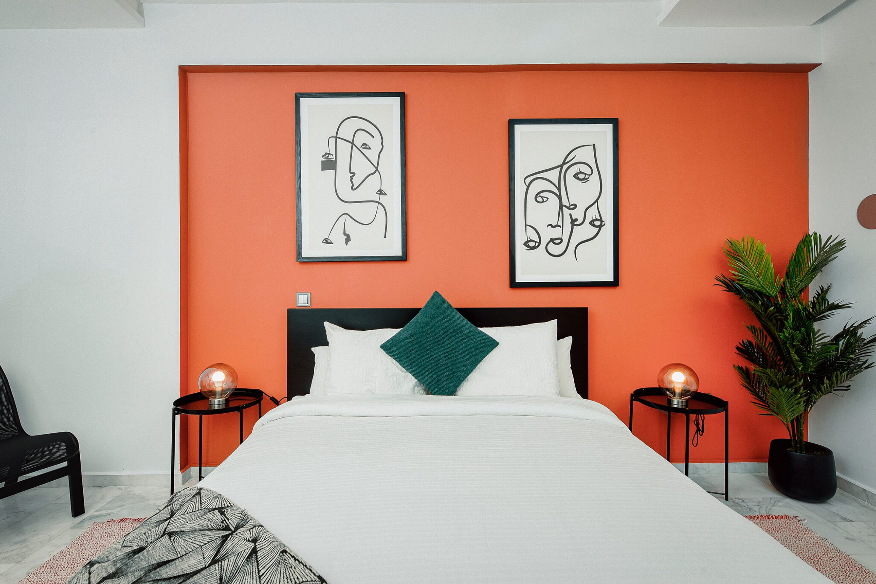 Un dormitorio moderno con una cama grande, una pared de acento naranja, dos cuadros enmarcados, un cojín verde y una planta en maceta.