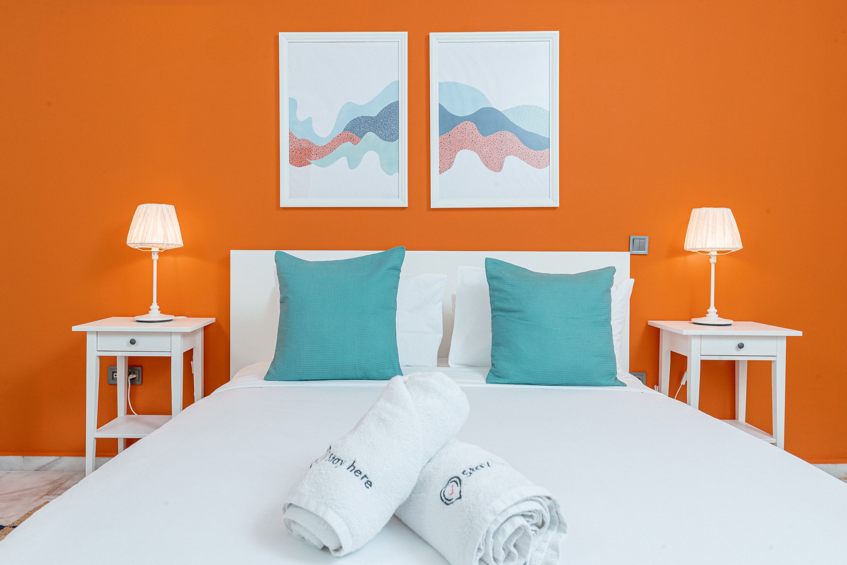 Un letto ben rifatto con lenzuola bianche e cuscini color acquamarina, affiancato da due comodini bianchi con lampade, su uno sfondo di parete arancione con due opere d'arte astratte.