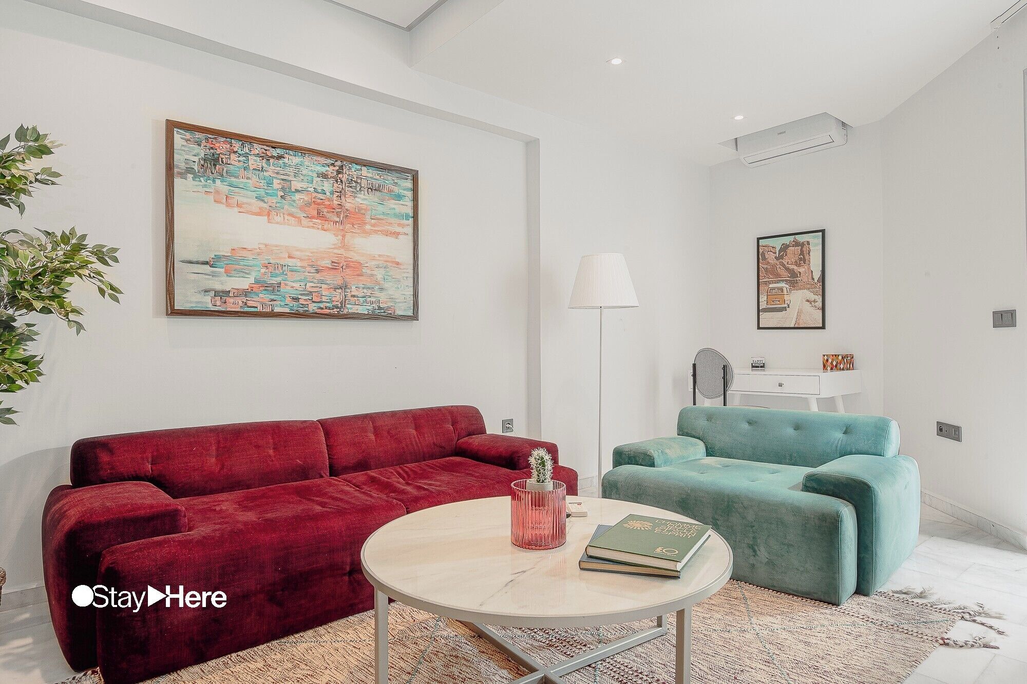 A modern living room with a red sofa, a teal armchair, and a round coffee table. There are two paintings on the walls and a floor lamp.