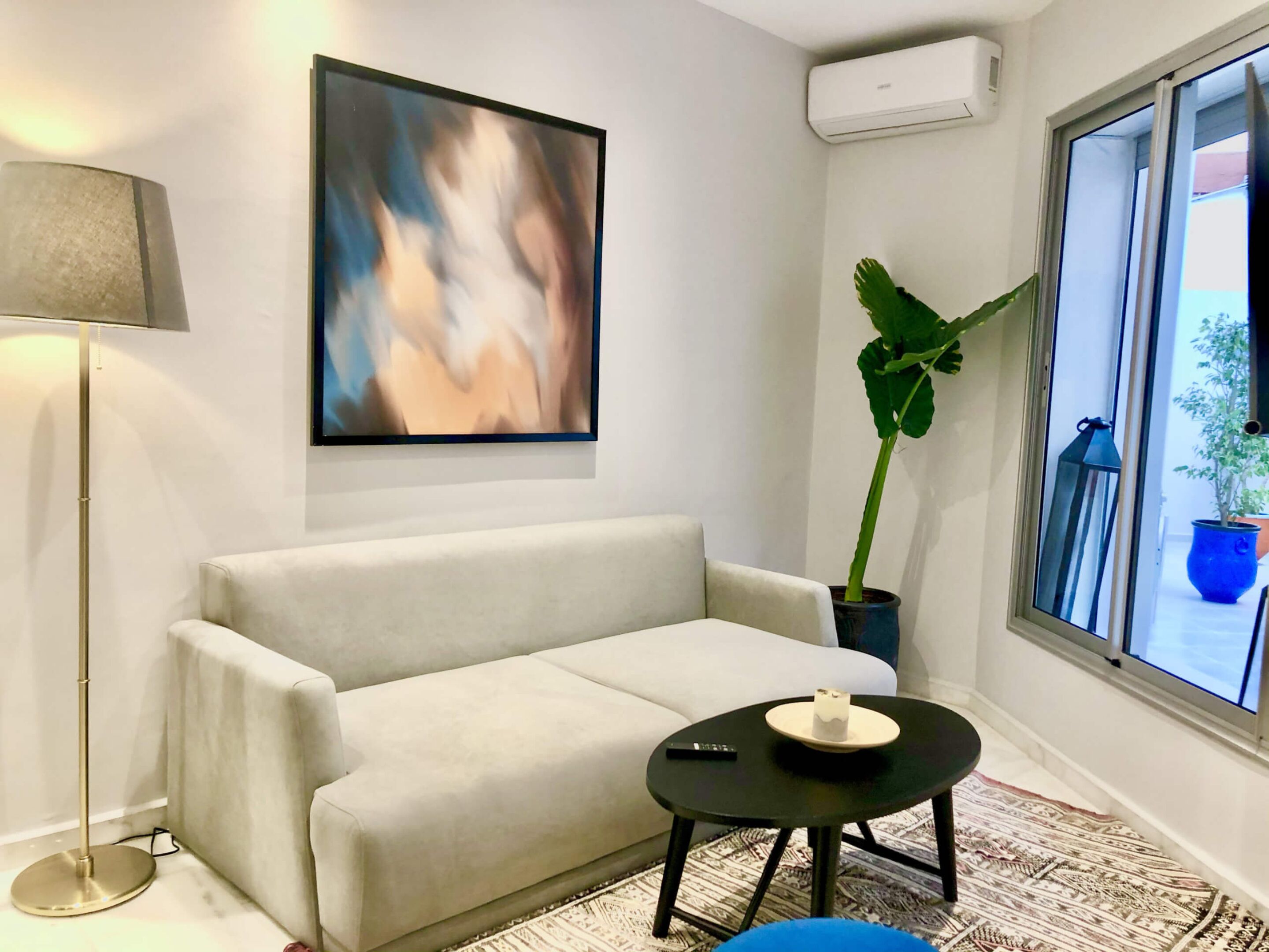 A modern living room with a sofa, a small black table with a remote control, a large abstract painting, a potted plant, and a window with a view.