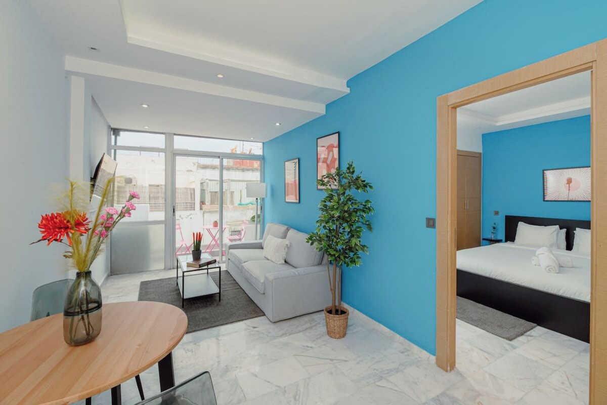 Una habitación de hotel moderna con una pared azul de acento, una cama grande, un sofá, una mesa de comedor con un jarrón de flores y vistas a través de una puerta de cristal.