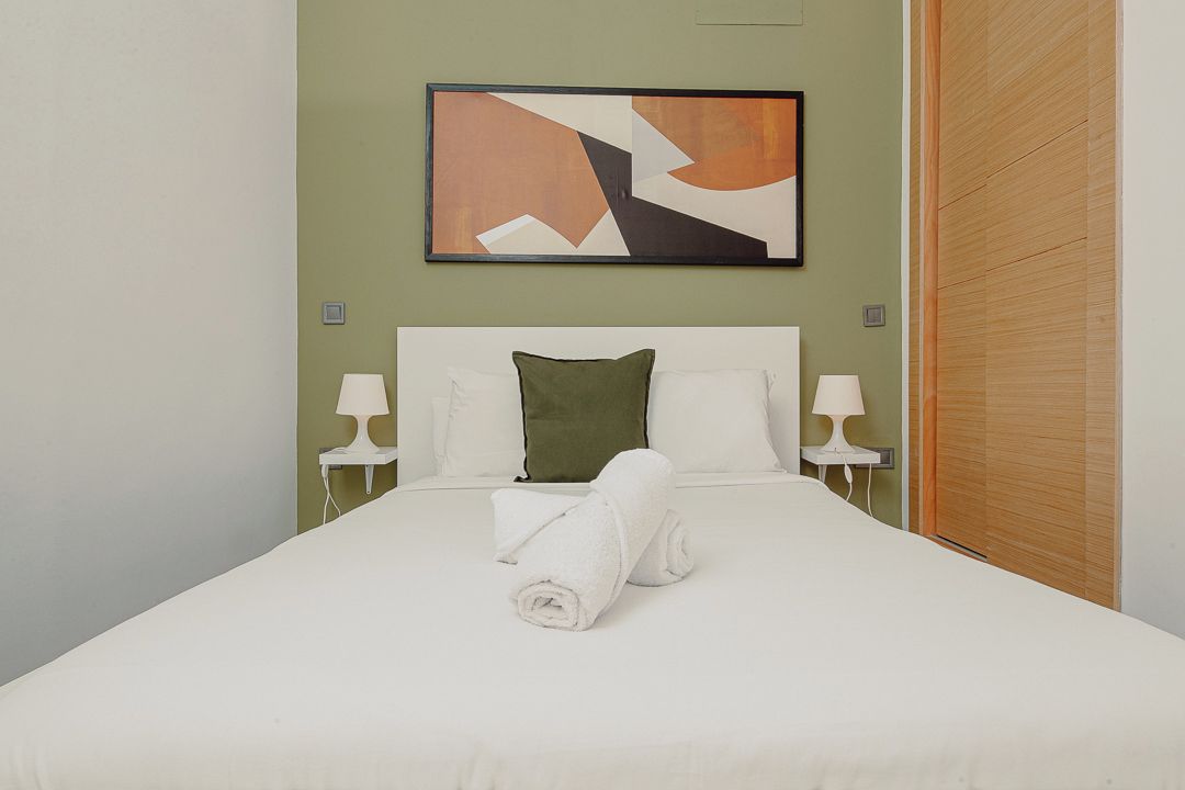 A hotel room with a bed, two bedside lamps, a green pillow, a white towel, and a painting on the wall.