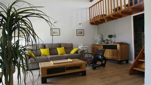 A living room with a wooden floor, a sofa with yellow cushions, a coffee table, a chair, a plant, a staircase, and a desk with a fan. - 3 bedroom house, private pool, 10min from Pont du Gard (Meynes)