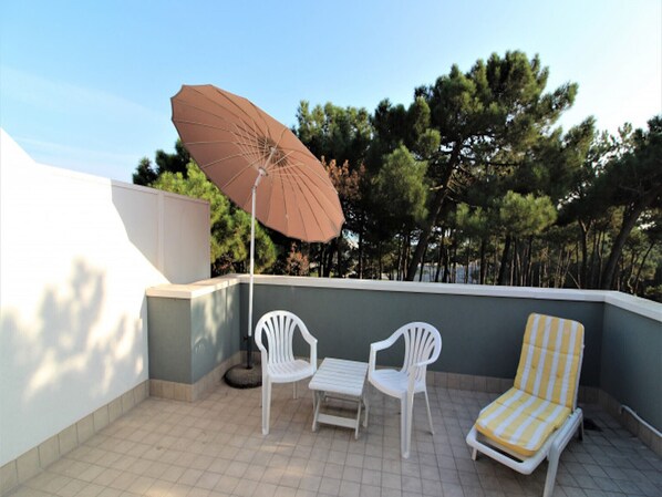 A balcony with two white chairs, a striped lounge chair, and a brown umbrella.