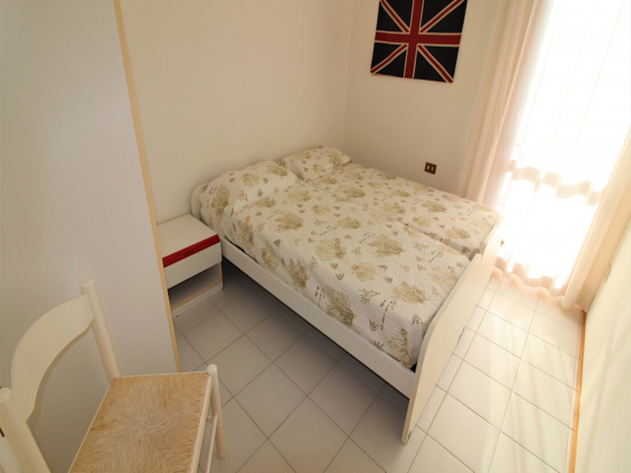 A small, neatly arranged bedroom with a single bed, a chair, and a wall-mounted flag.