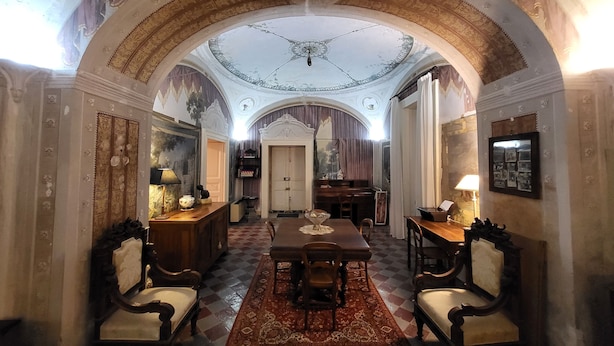 Un salón imponente con un techo abovedado alto, decoraciones murales elaboradas y una mesa central con sillas.