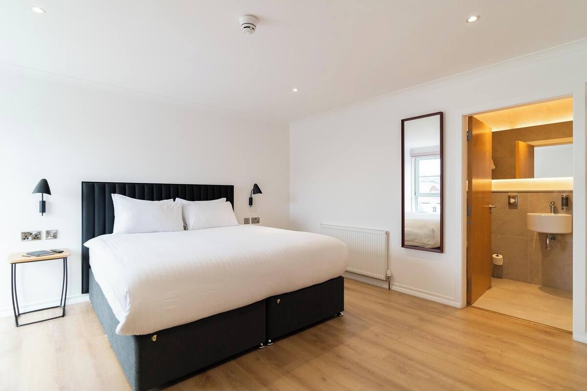 A modern hotel room with a large bed, bedside lamps, a radiator, and a bathroom with a sink and mirror.