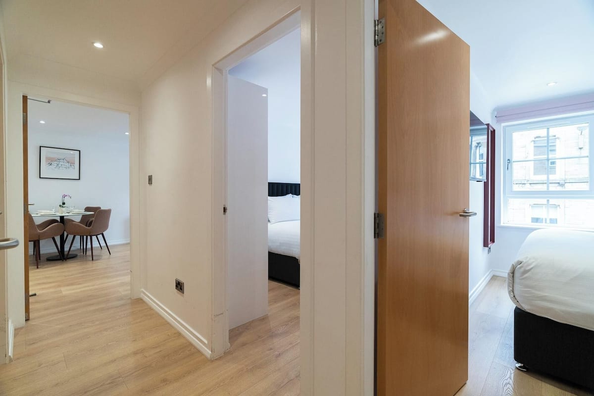 A modern hotel room with a wooden door, a bed, a window with a city view, and a dining area with a table and chairs.