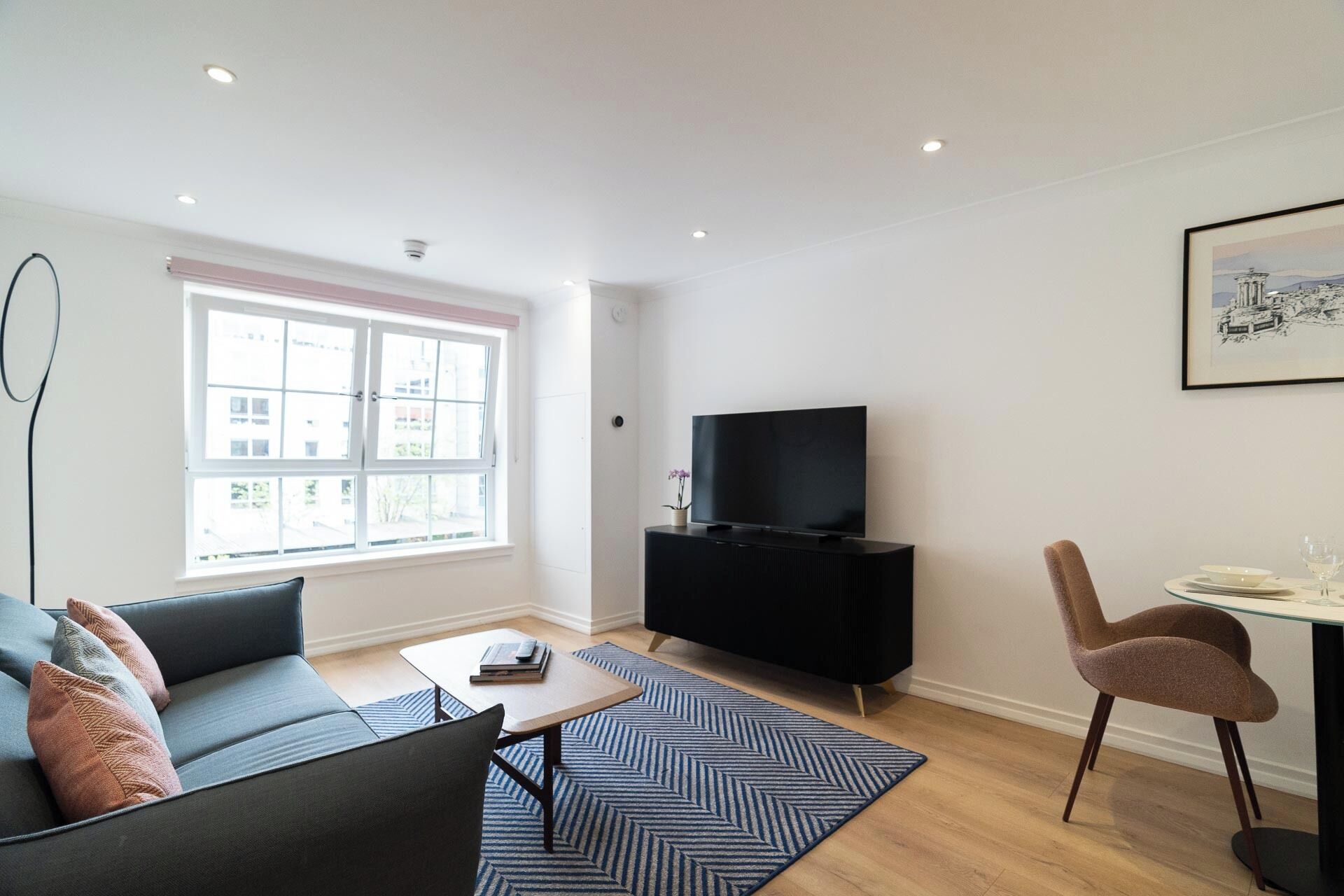 A modern living room with a sofa, a coffee table, a TV on a black stand, a dining table with a chair, and a large window with blinds.