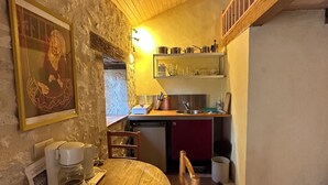 Una cocina acogedora con una mesa de madera, sillas y una cafetera. Hay un lavadero, una estufa y un estante con utensilios de cocina.