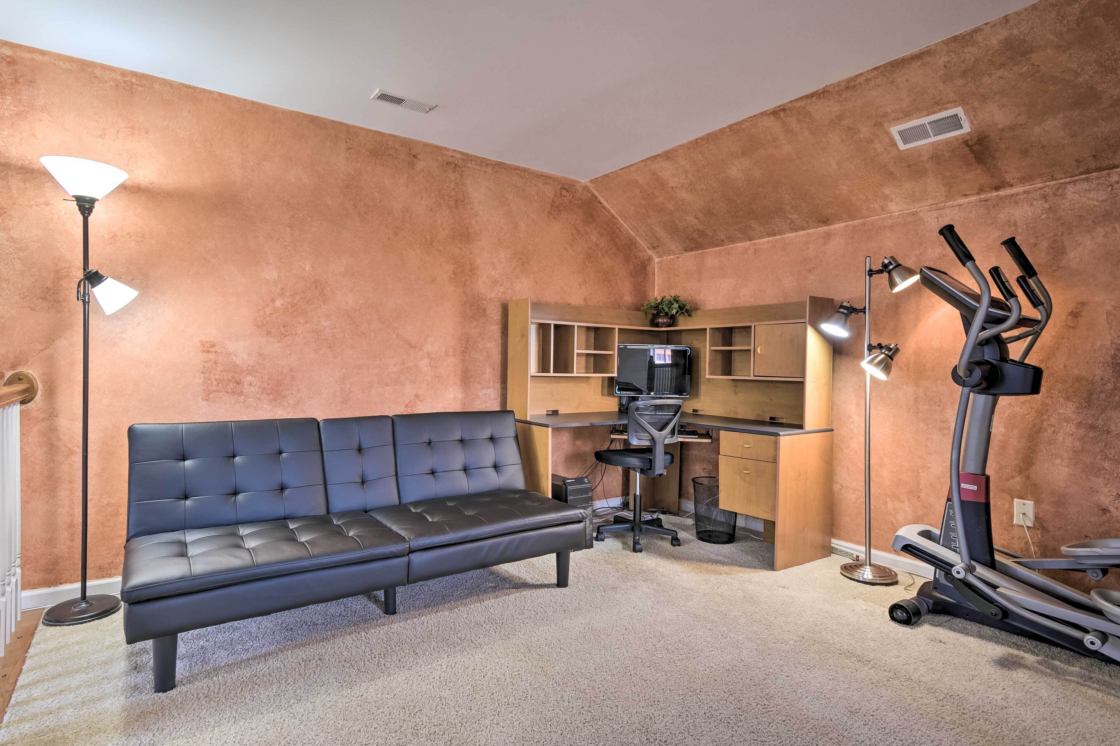 A room with a sofa, desk, chair, treadmill, and two floor lamps.