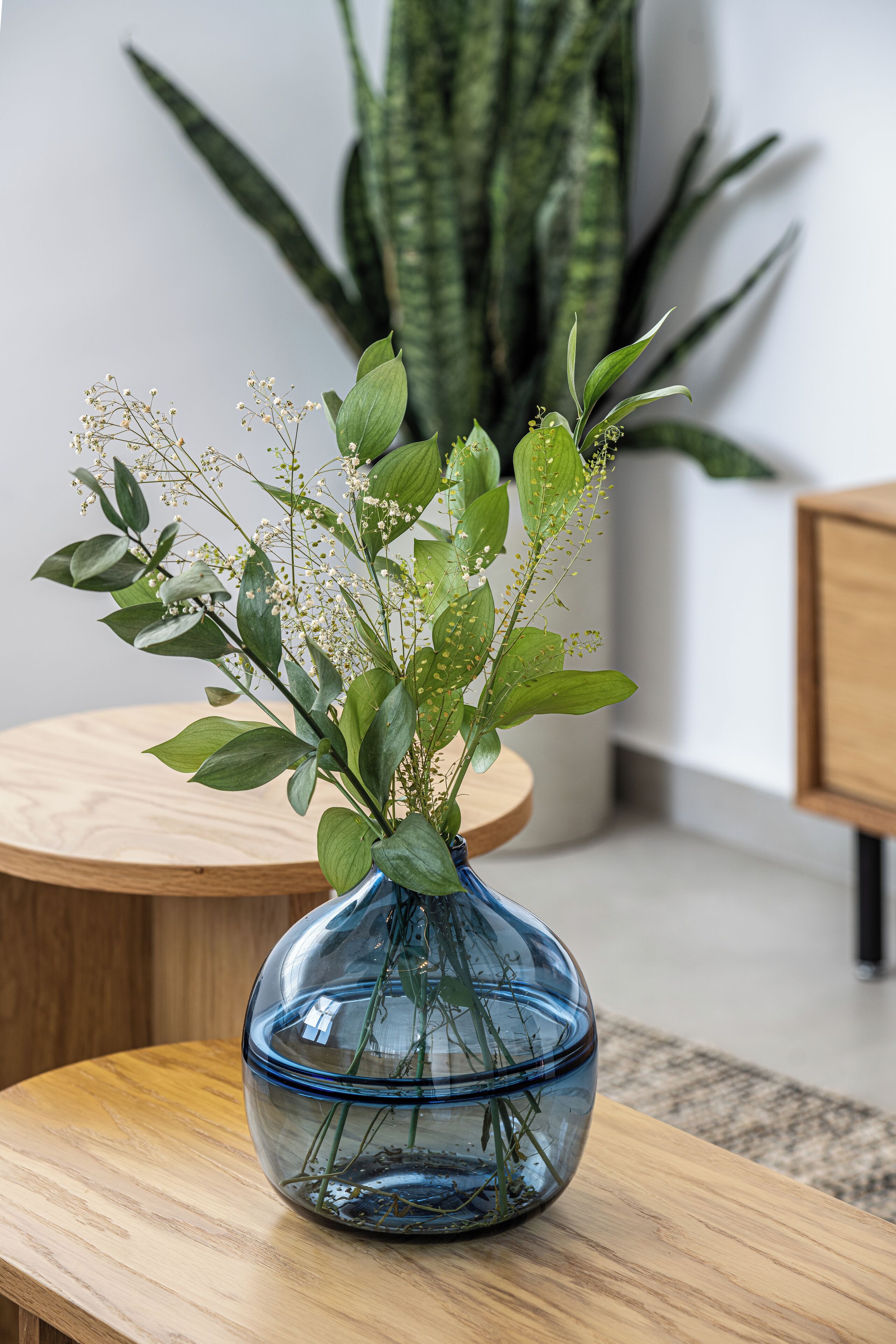 A blue glass vase with green foliage and small white flowers on a wooden table.