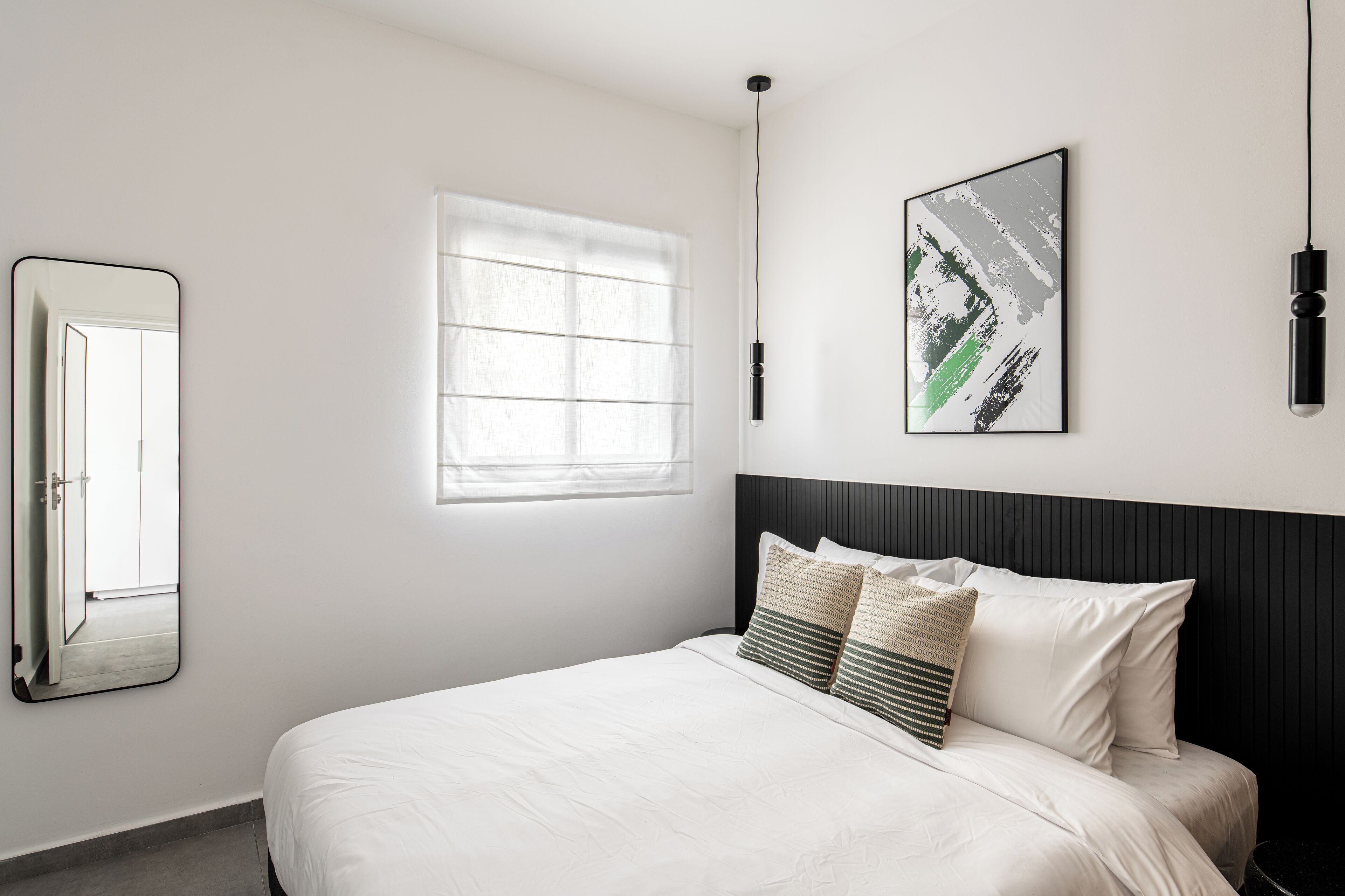 A neatly arranged bedroom with a bed, a window, a wall-mounted mirror, and a framed artwork.