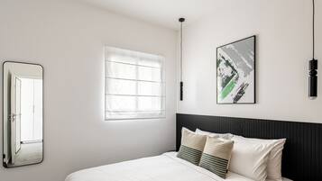 A neatly arranged bedroom with a bed, a window, a wall-mounted mirror, and a framed artwork.