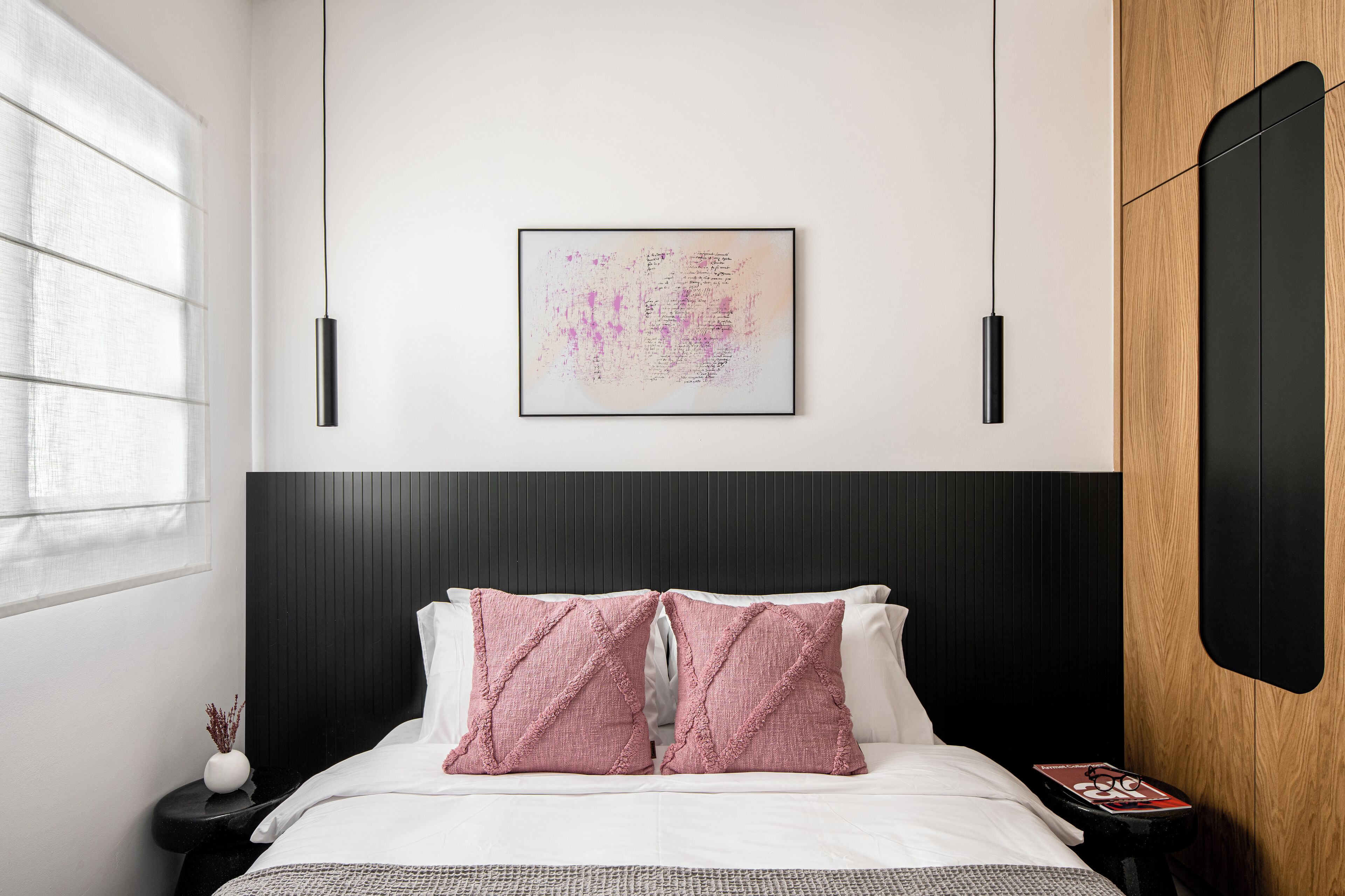 Une chambre moderne avec une tête de lit noire, du linge de lit blanc et des coussins roses. Un tableau encadré est accroché au mur, et une petite table porte un livre et des lunettes.