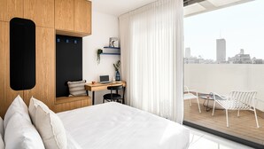 A modern hotel room with a large bed, a desk with a laptop, a small seating area with chairs, and a view of the city through a large window. - Mate GEORGE - By HOMY (Tel Aviv)