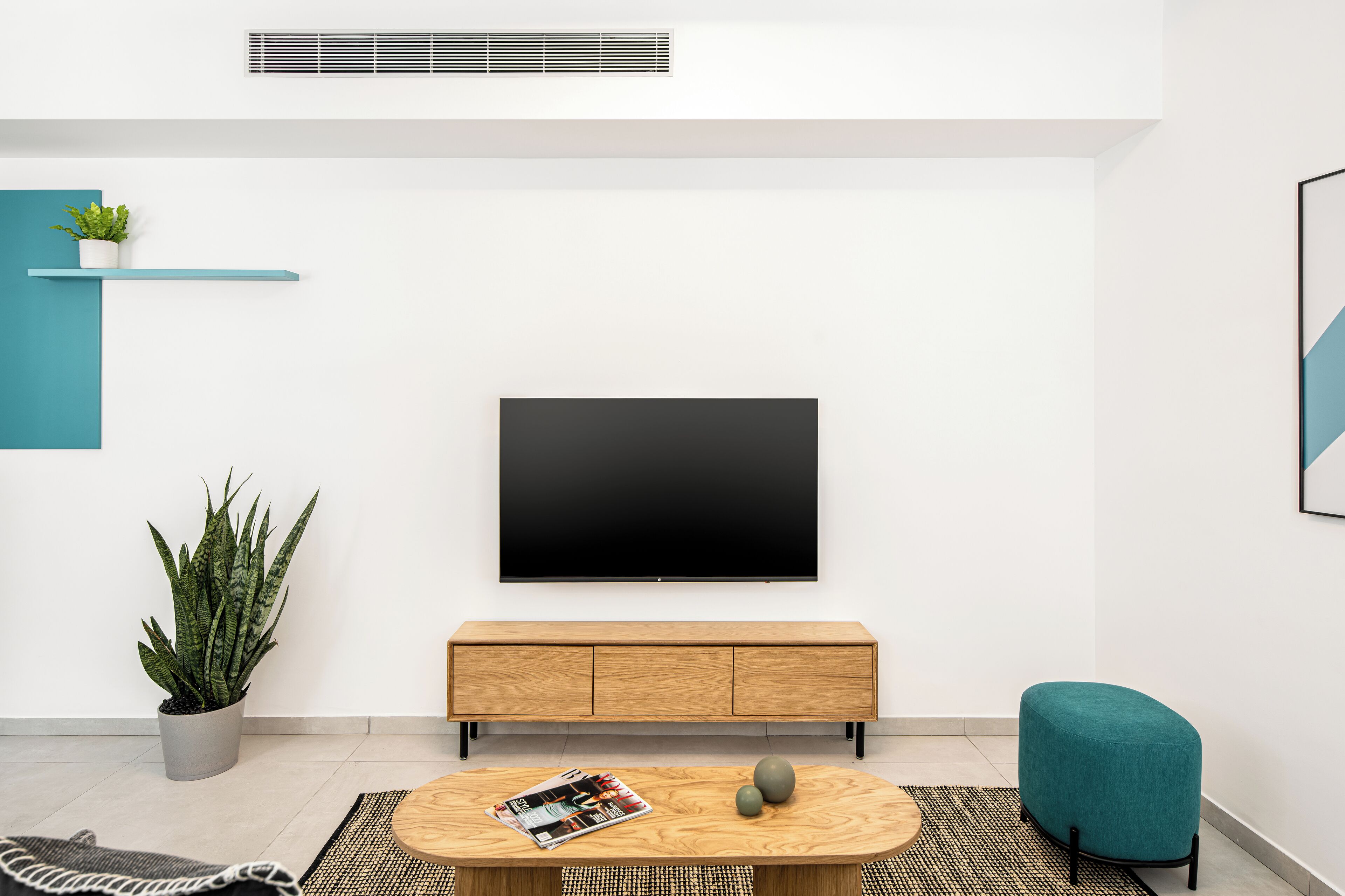 Una sala moderna con un televisor de pantalla plana montado en la pared, una consola de madera, un otoman turquesa y dos plantas en macetas.