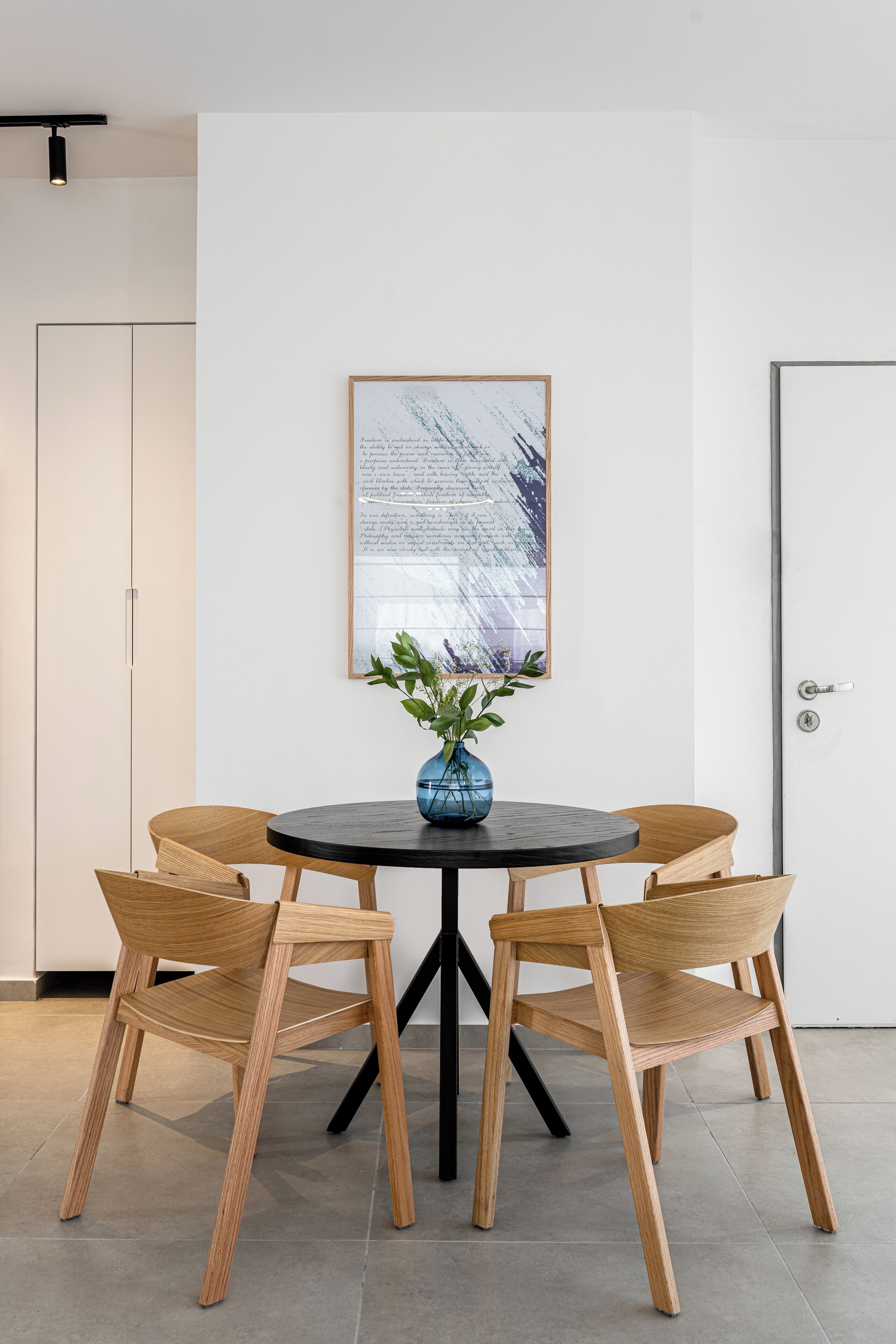 A dining area with a round table, four wooden chairs, a blue vase with greenery, and a framed abstract painting on the wall.
