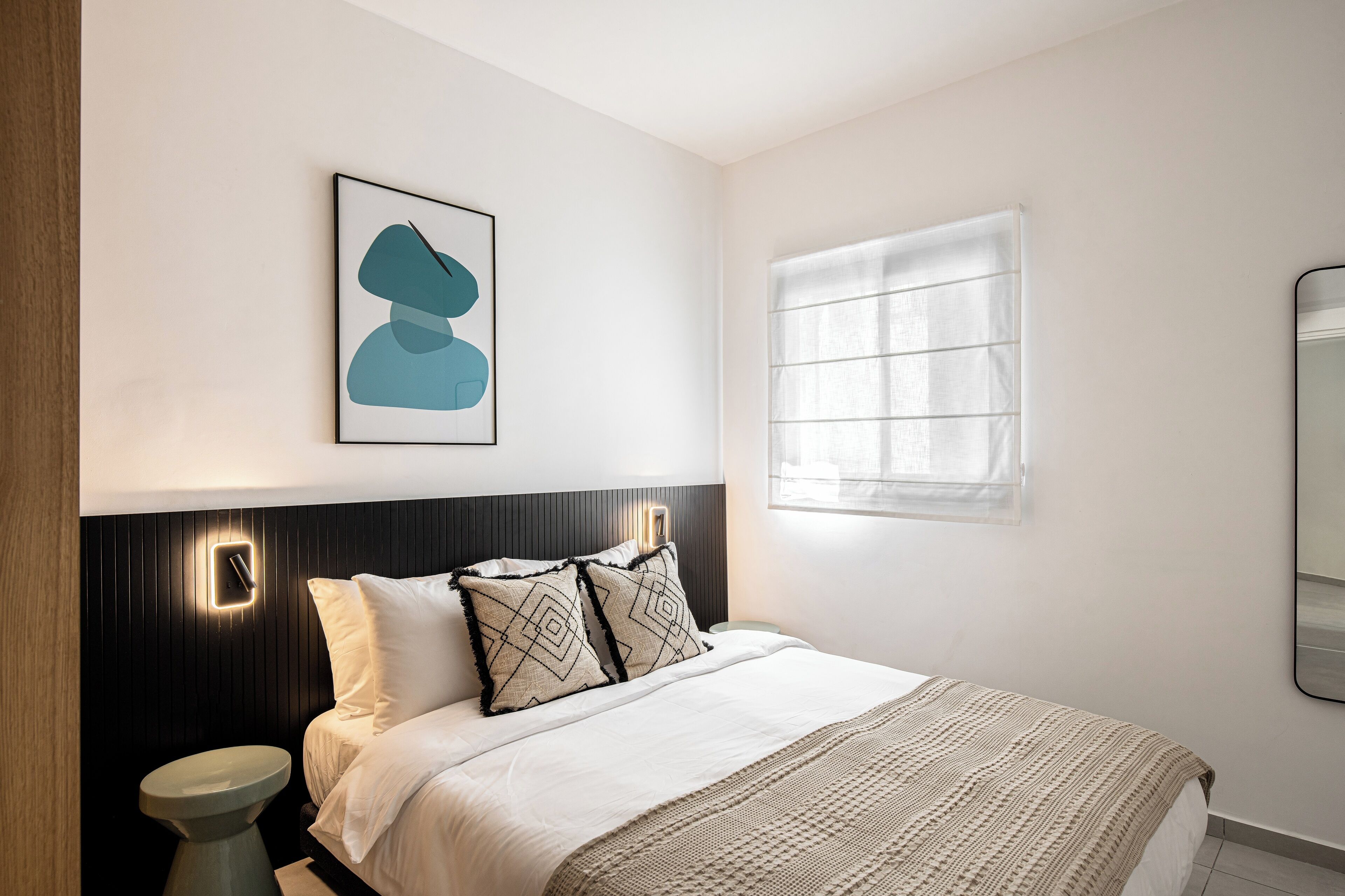 A modern bedroom with a bed, a bedside table, a wall-mounted light, a window with blinds, and a framed abstract artwork.