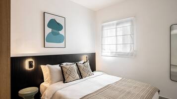 A modern bedroom with a bed, a bedside table, a wall-mounted light, a window with blinds, and a framed abstract artwork.