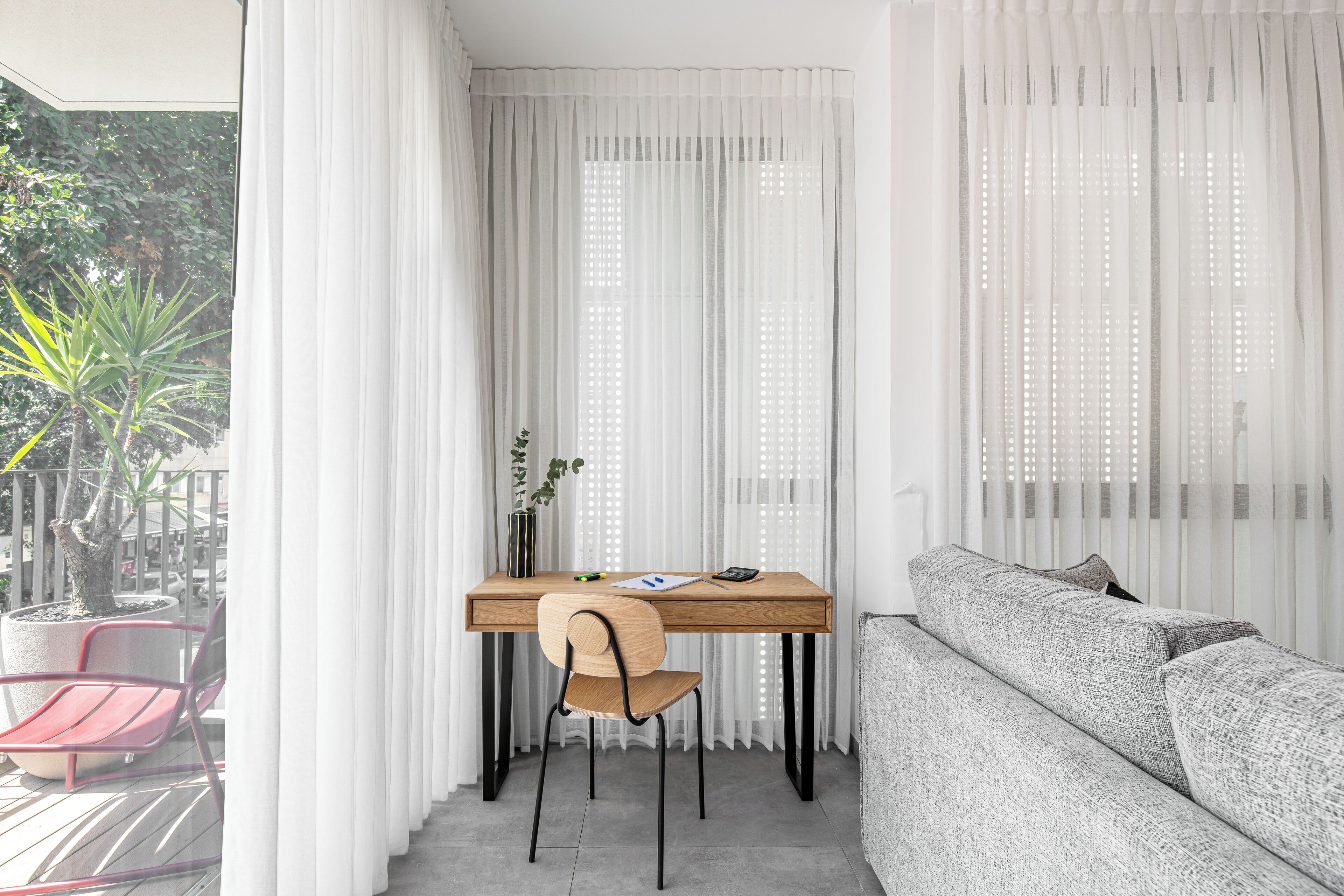Una habitación moderna con un escritorio de madera, una silla, un sofá gris y cortinas blancas.