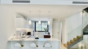 Eine moderne Küche mit einer zentralen Kochinsel, Barhocker, Pendelleuchten und Blick ins Freie durch ein Fenster.