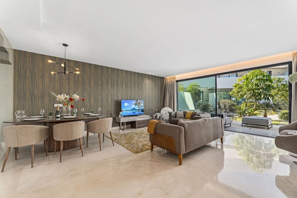 A modern living room with a dining area, a flat-screen TV, and a large window overlooking a garden.