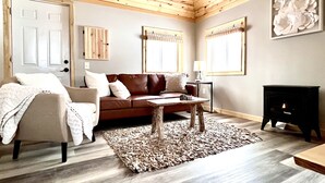 Een gezellige woonkamer met een houten plafond, een bruine leren bank, een beige fauteuil, een salontafel en een zwarte open haard.